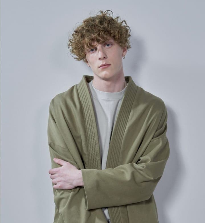 A curly-haired young man in an olive jacket, arms crossed