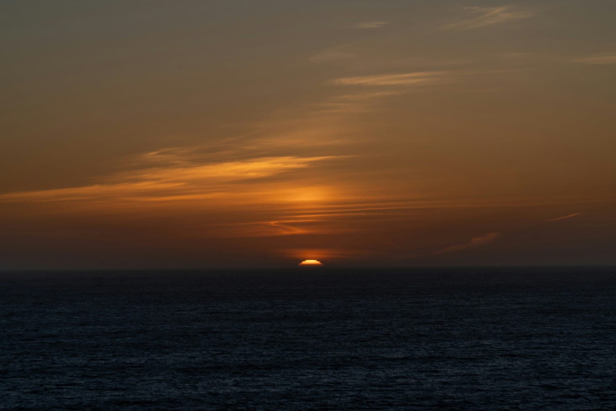 The sun setting over a dark sea under streaks of cloud